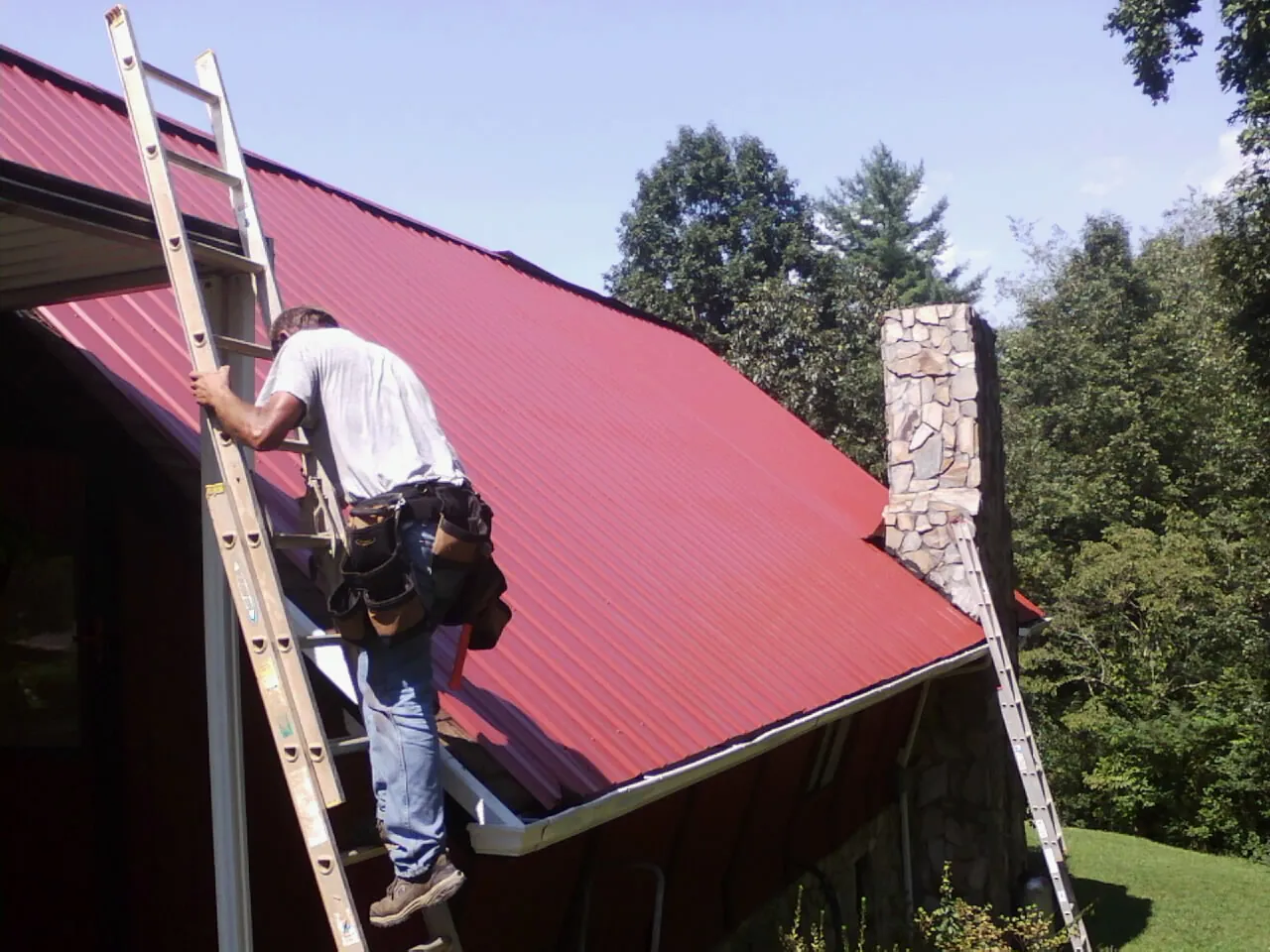 Roofer on a ladder beside a red metal roof and stone chimney — Soffit & Fascia Repair by Roofing Brothers in Charlotte Hall